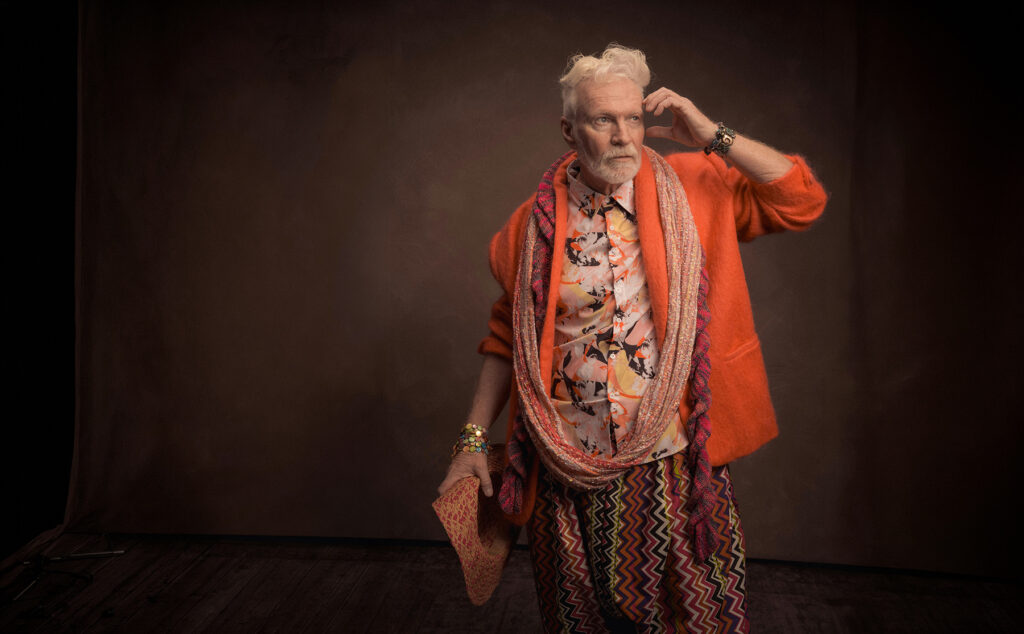 Homem sênior estiloso com roupas coloridas e texturizadas, posando para um retrato contra fundo neutro.